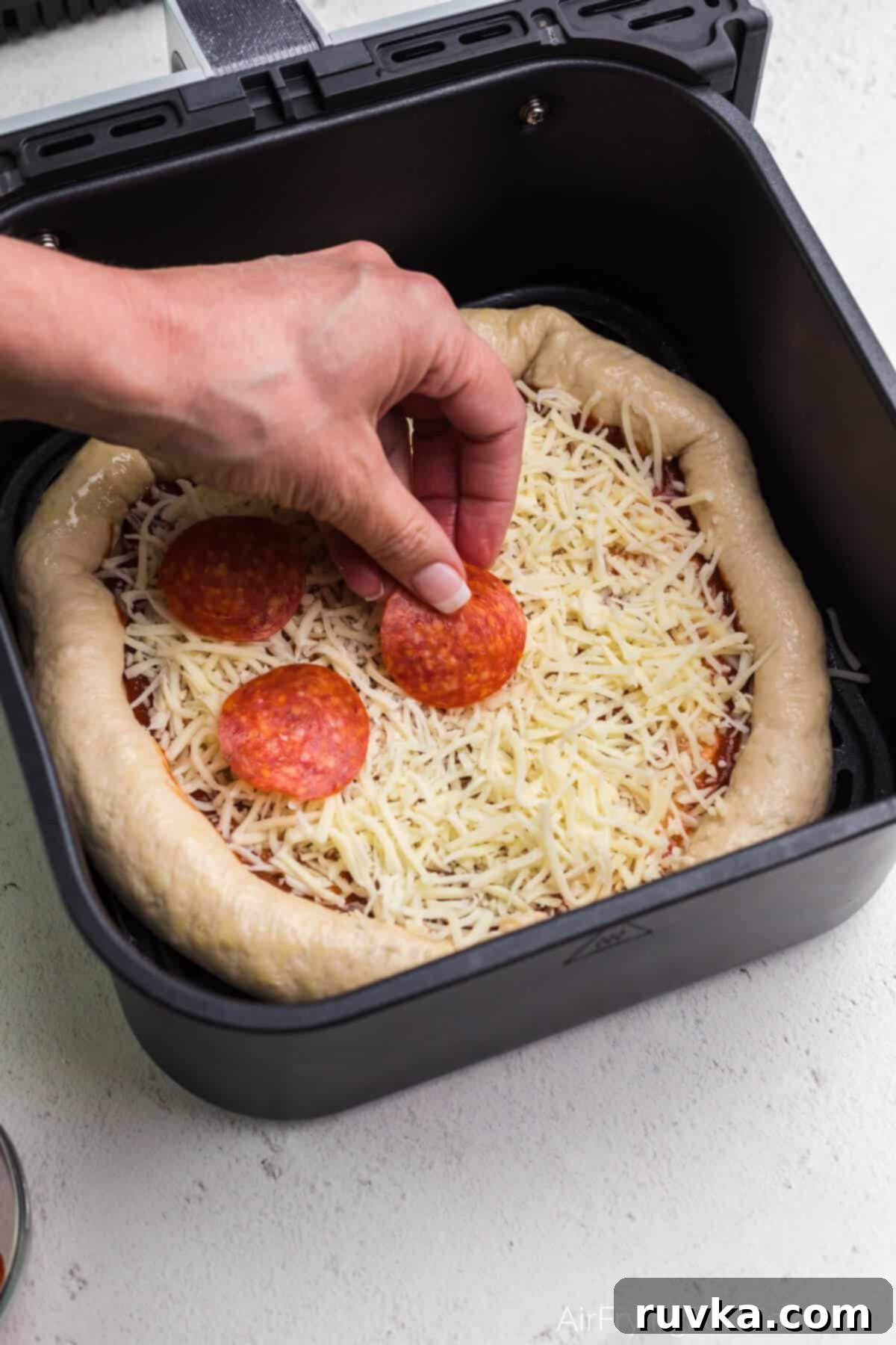 Crispy Air Fryer Pizza 7 Pepperoni slices being carefully arranged on top of the cheese layer on a pizza in an air fryer basket.