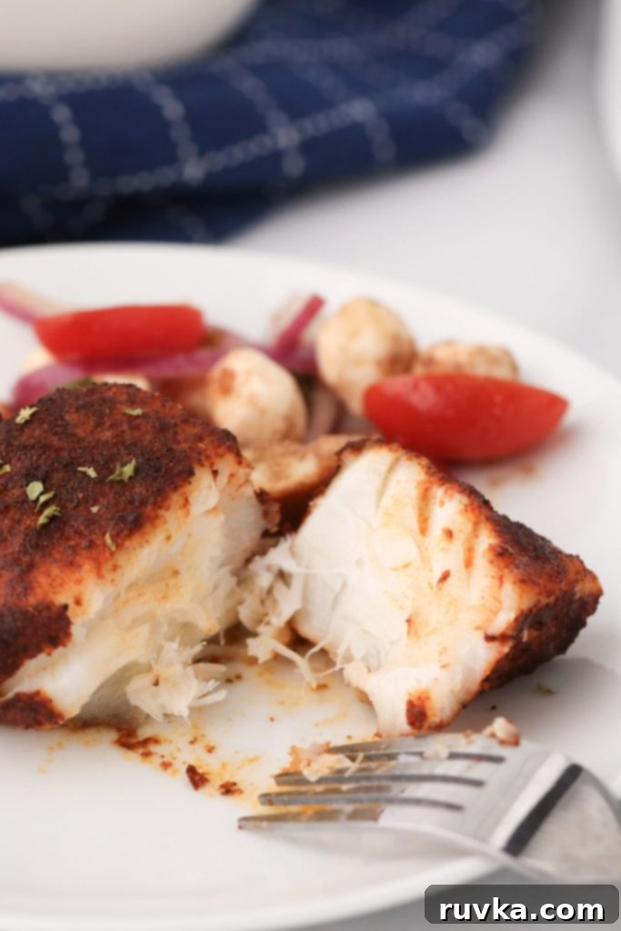 Perfectly Flaky Air Fryer Cod 7 Flaky cod seasoned and broken in half with a fork on the plate, garnished with tomatoes and vegetables, showing the tender interior