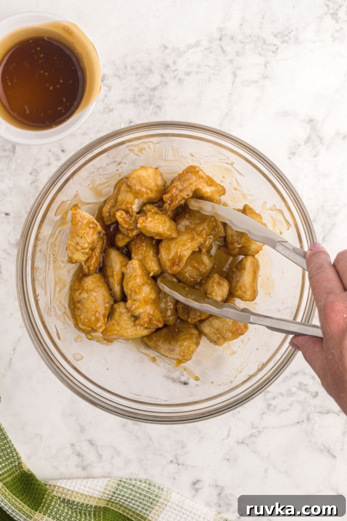 Crispy Air Fryer Sweet & Tangy Chicken 8 Tossing cooked chicken in a clear glass bowl with sweet and sour sauce on a white marble table