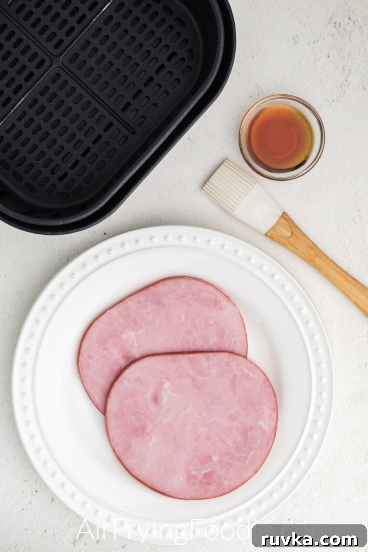 Easy Air Fryer Ham Steak 3 ham steaks and maple syrup with a silicone basting brush on a white table.