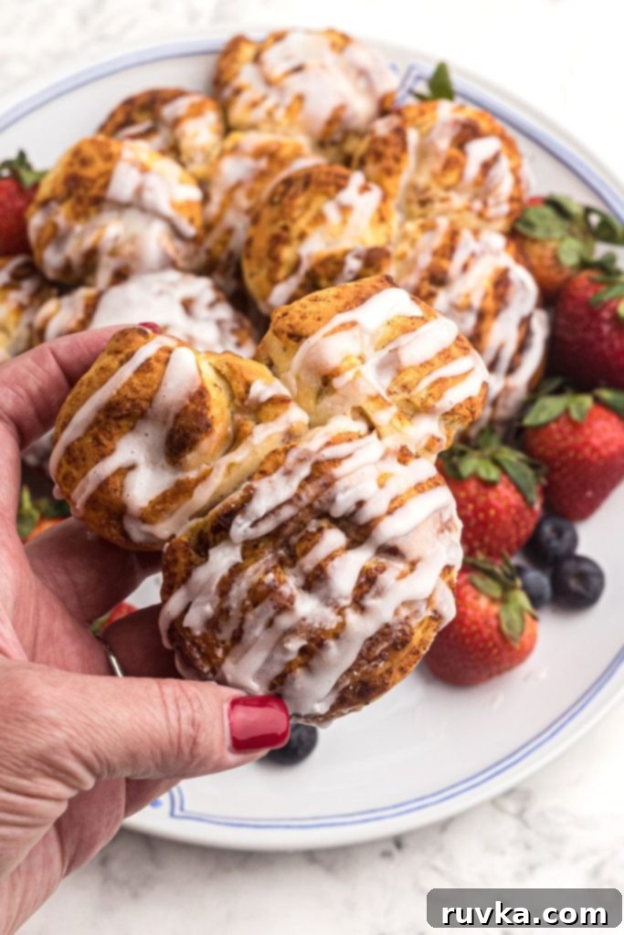 Mickey's Air Fryer Cinnamon Rolls 9 A hand holding a perfectly frosted, golden-brown Mickey Mouse shaped cinnamon roll, with a plate of fresh fruit in the background.
