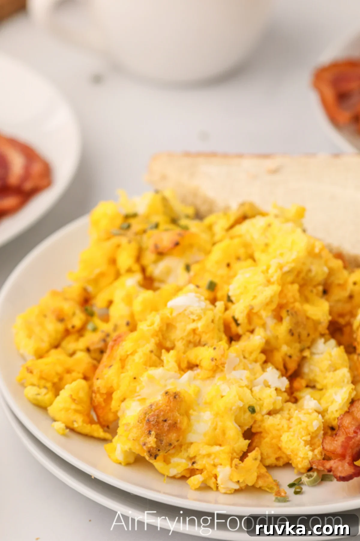 Fluffy Air Fryer Scrambled Eggs 2 Scrambled eggs that were made in the air fryer on a plate with bacon and toast.