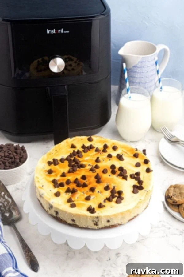 Chocolate Chip Cookie Cheesecake in the Air Fryer 5 A whole, perfectly cooked air fryer chocolate chip cheesecake, served on an elegant white cake stand, positioned in front of an air fryer, ready to be sliced and enjoyed.