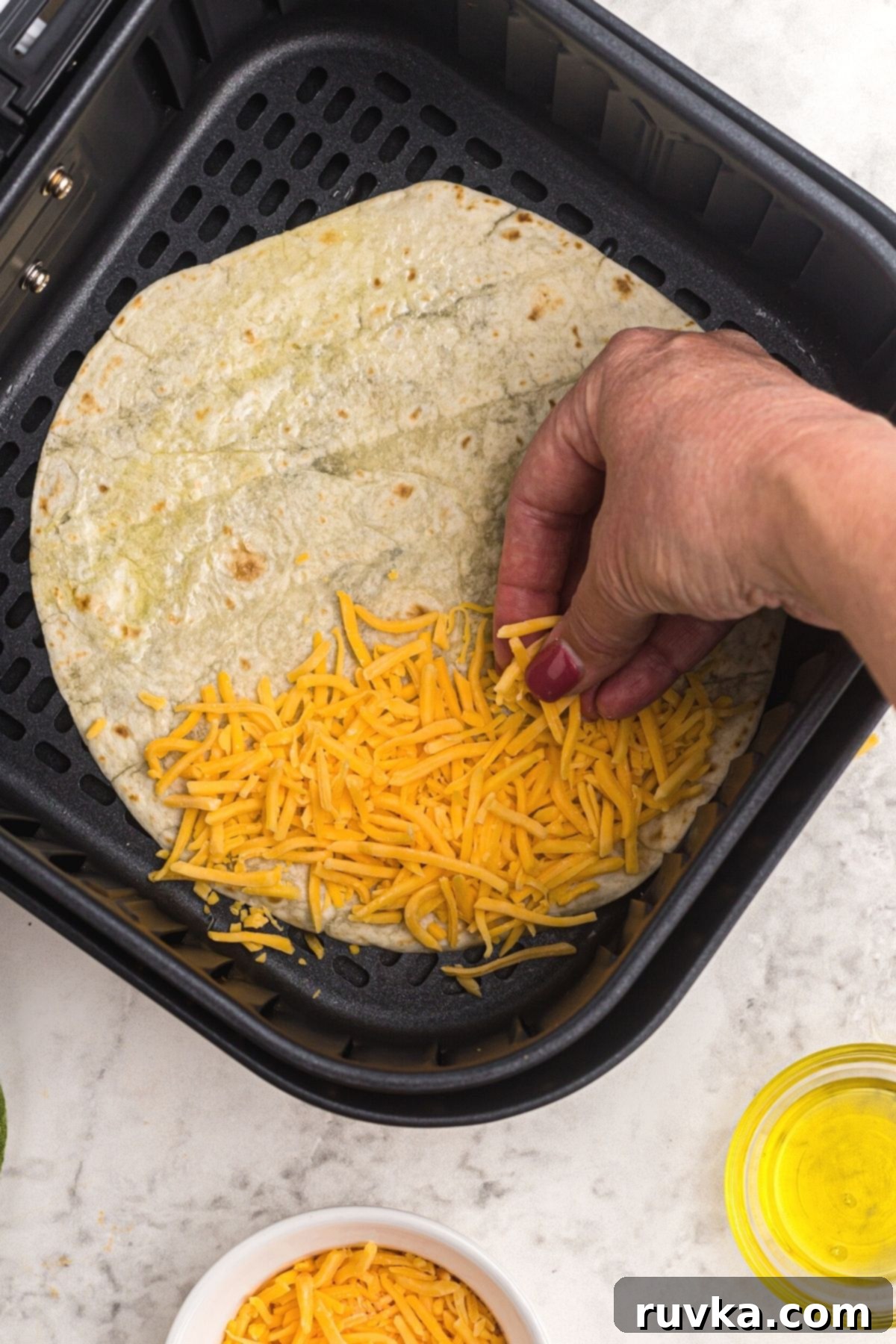 Crispy Air Fryer Quesadillas 5 A hand sprinkling an even layer of shredded cheddar cheese onto a flour tortilla in the air fryer basket, preparing it for folding.