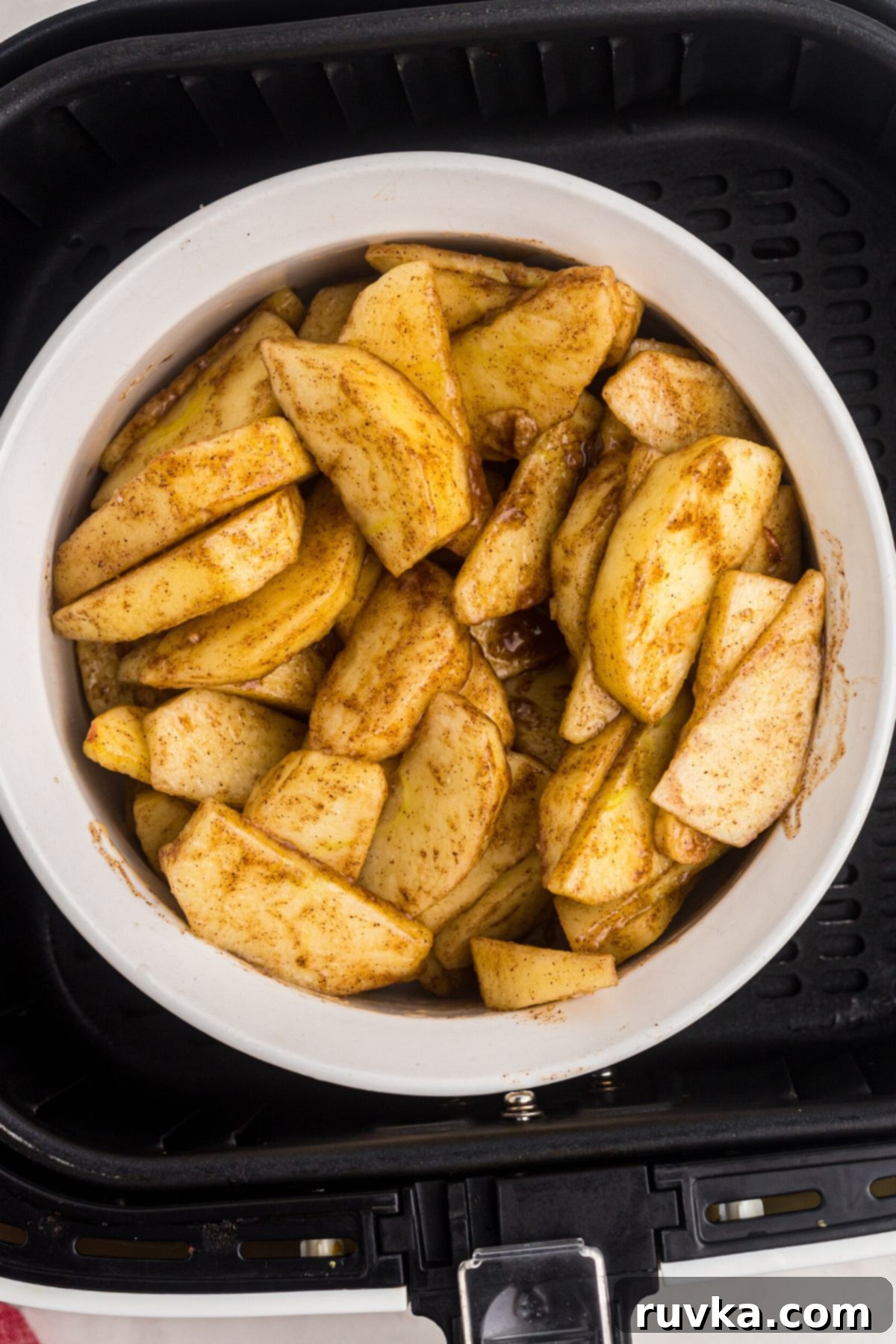 Perfect Air Fryer Apples 5 Sliced apples, glistening with a perfect coating of cinnamon and brown sugar, mixed in a white Pyrex bowl, now placed inside the air fryer basket, ready to be cooked to tender perfection.