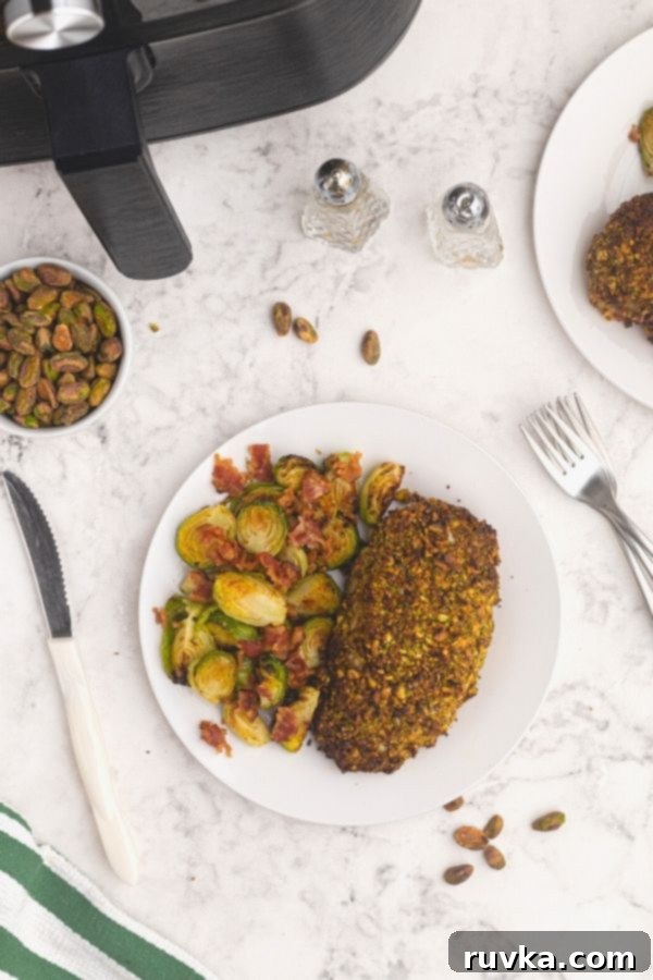 Crispy Pistachio Air Fryer Chicken 3 Pistachio crusted chicken cut into slices, served on a white plate next to Brussel sprouts. In front of an air fryer, with pistachios scattered on the table.