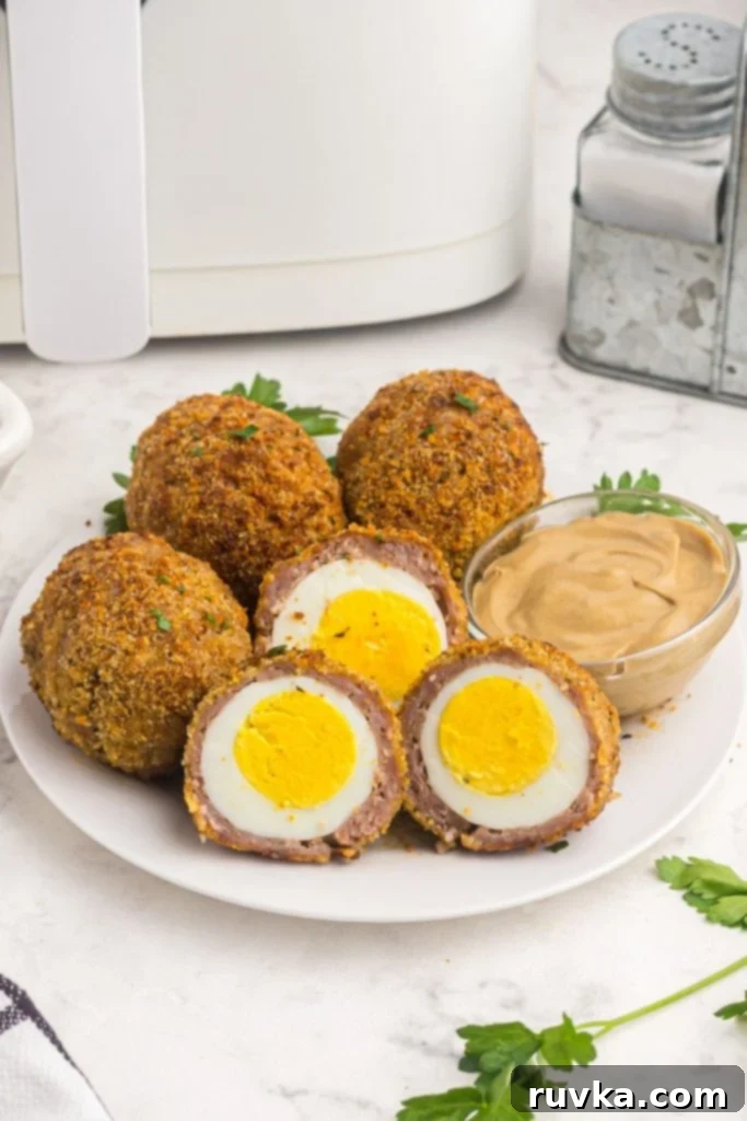 The Best Air Fryer Scotch Eggs 2 Coated hard boiled eggs with sausage and breadcrumbs, served on a white plate with Dijon mustard.