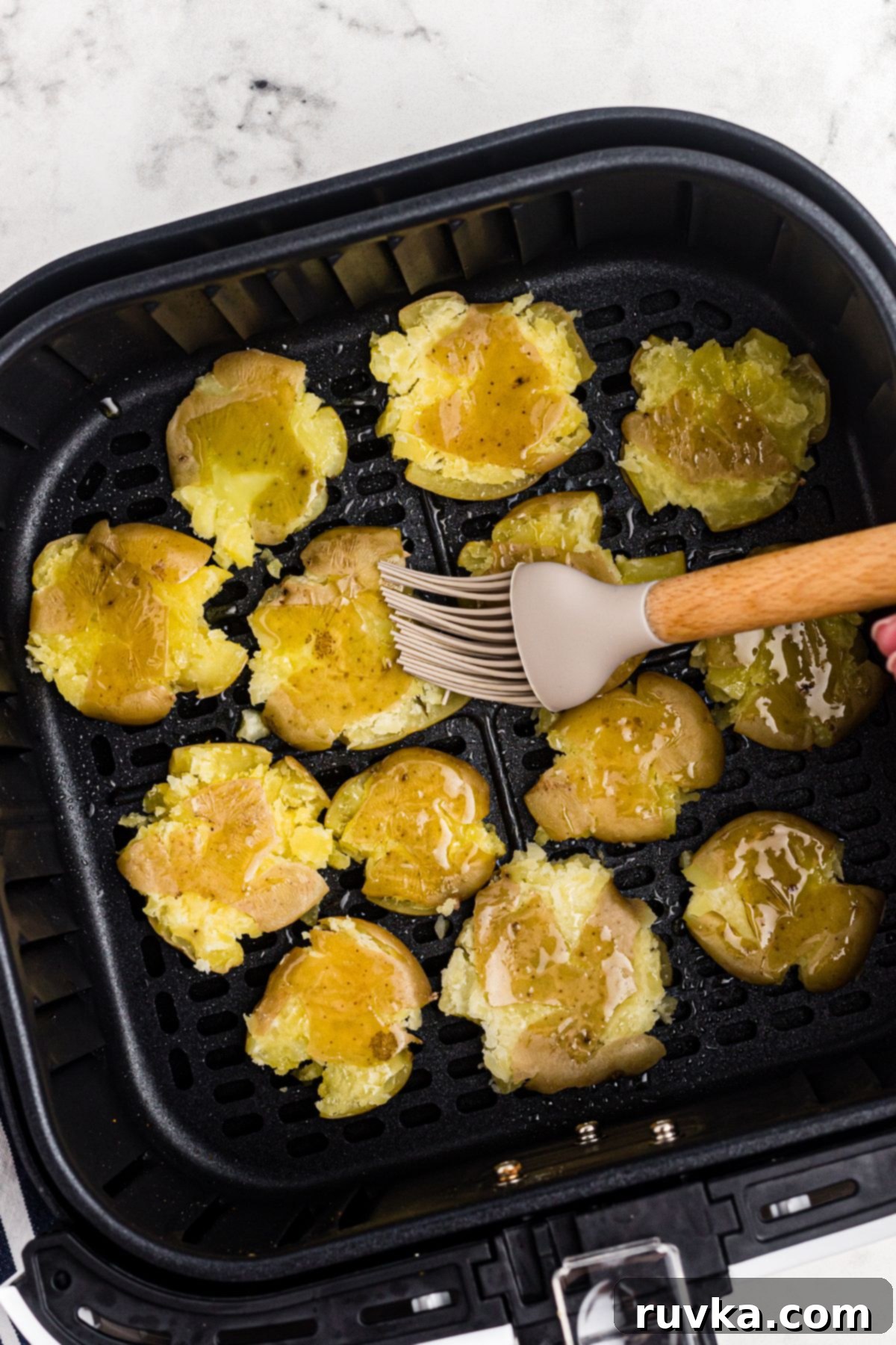 Air Fryer Crispy Smashed Potatoes 6 Smashed golden baby potatoes in the air fryer basket being brushed with olive oil and seasoned with spices.