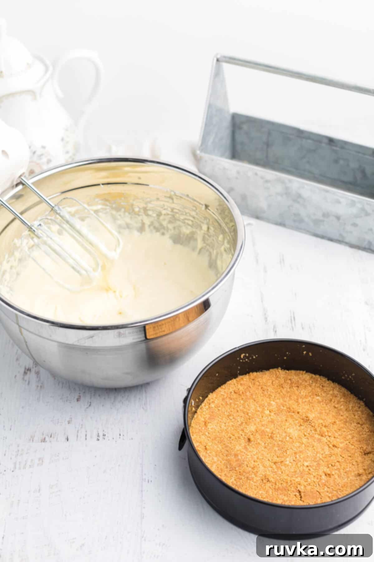 Perfect Air Fryer Cheesecake 4 Crushed graham crackers pressed into the bottom of a springform pan, with cheesecake filling ingredients in a mixing bowl alongside, ready to be poured.