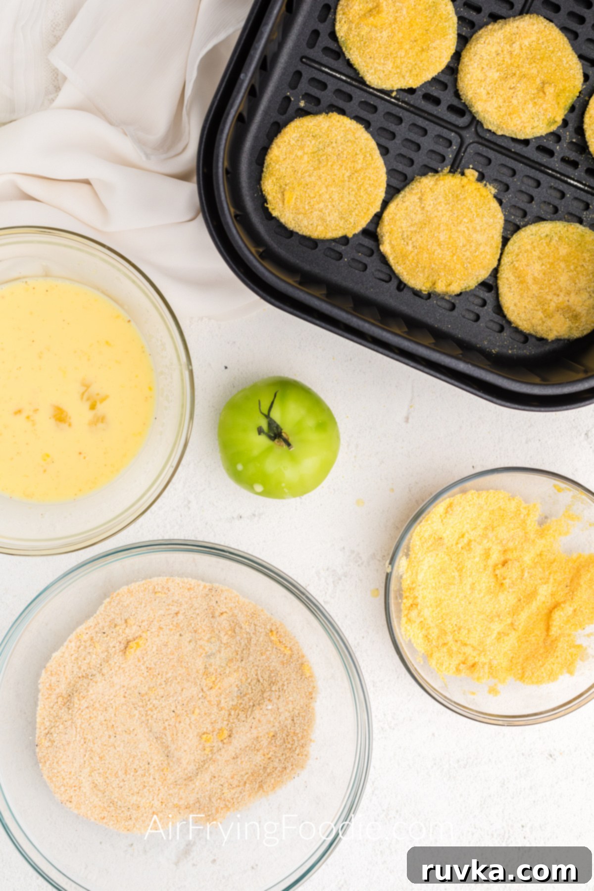 Crispy Air Fryer Fried Green Tomatoes 6 Coated green tomatoes in a single layer in the basket of the air fryer, surrouded by ingredients on a table.