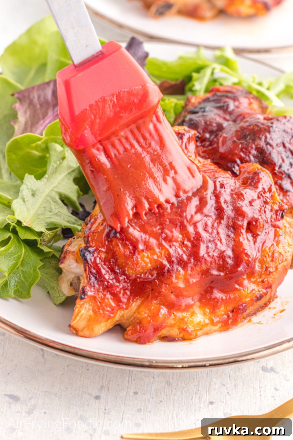 Perfect Air Fryer BBQ Chicken Thighs 4 BBQ chicken thighs being brushed with additional BBQ sauce on a plate with a side salad.